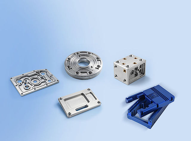 Mechanische Bauteile aus CNC-Fertigung: Dreh-, Fräs- und Stanzteile in verschiedenen Formen und Materialien, darunter Rundteile, Rechteckplatten und komplex gefräste Gehäuse auf hellblauem Hintergrund.